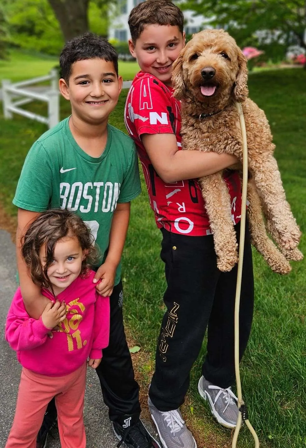 Three smiling children outdoors, the older boy holding their apricot Goldendoodle
